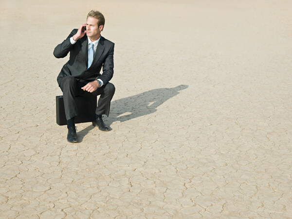 干旱地里坐着皮箱打手机的商务男士图片